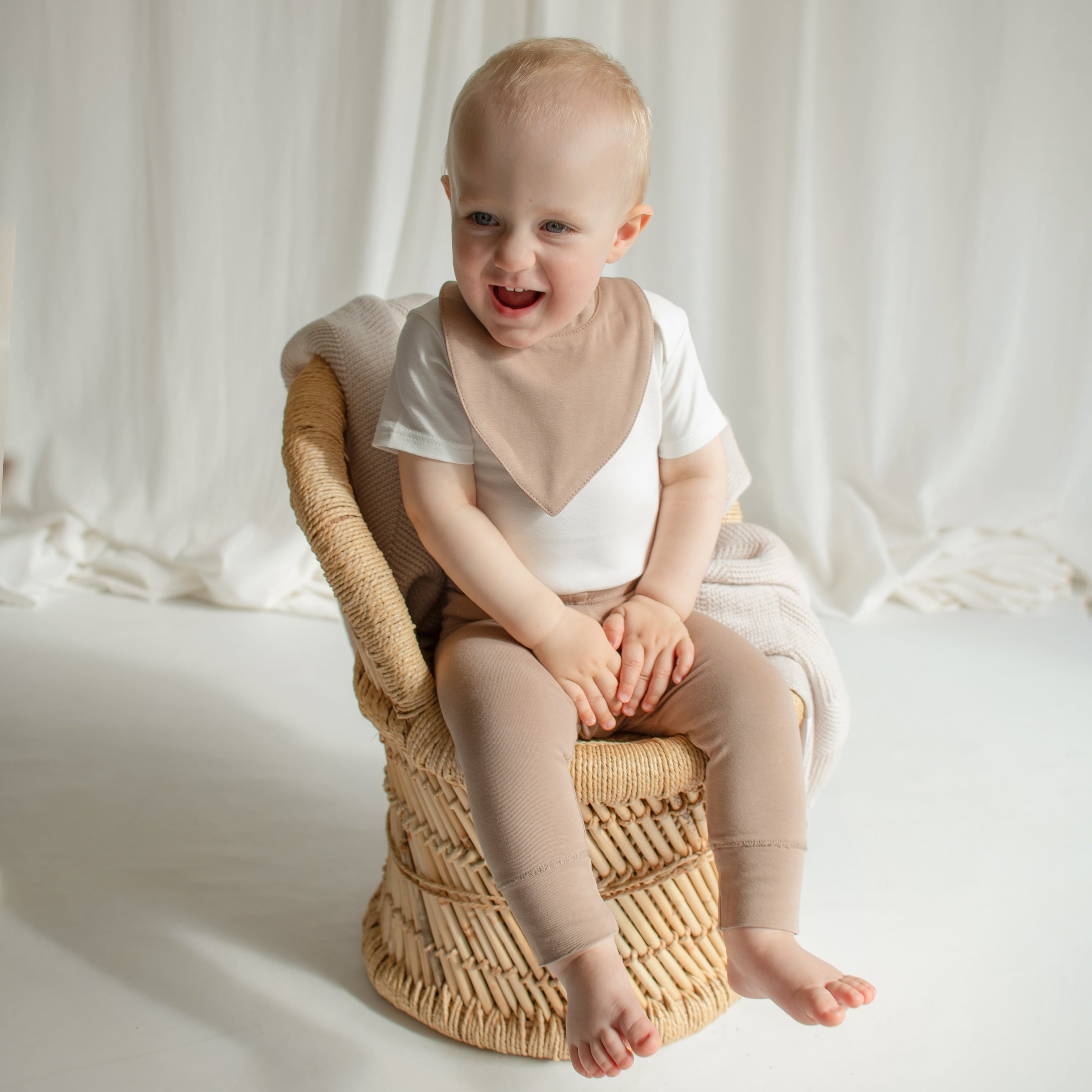 A little boy sitting on a small rattan chair, wearing a stylish CUUTTI outfit featuring an ivory short sleeve bodysuit, a cookie-colored waterproof bib, and cookie-colored leggings.